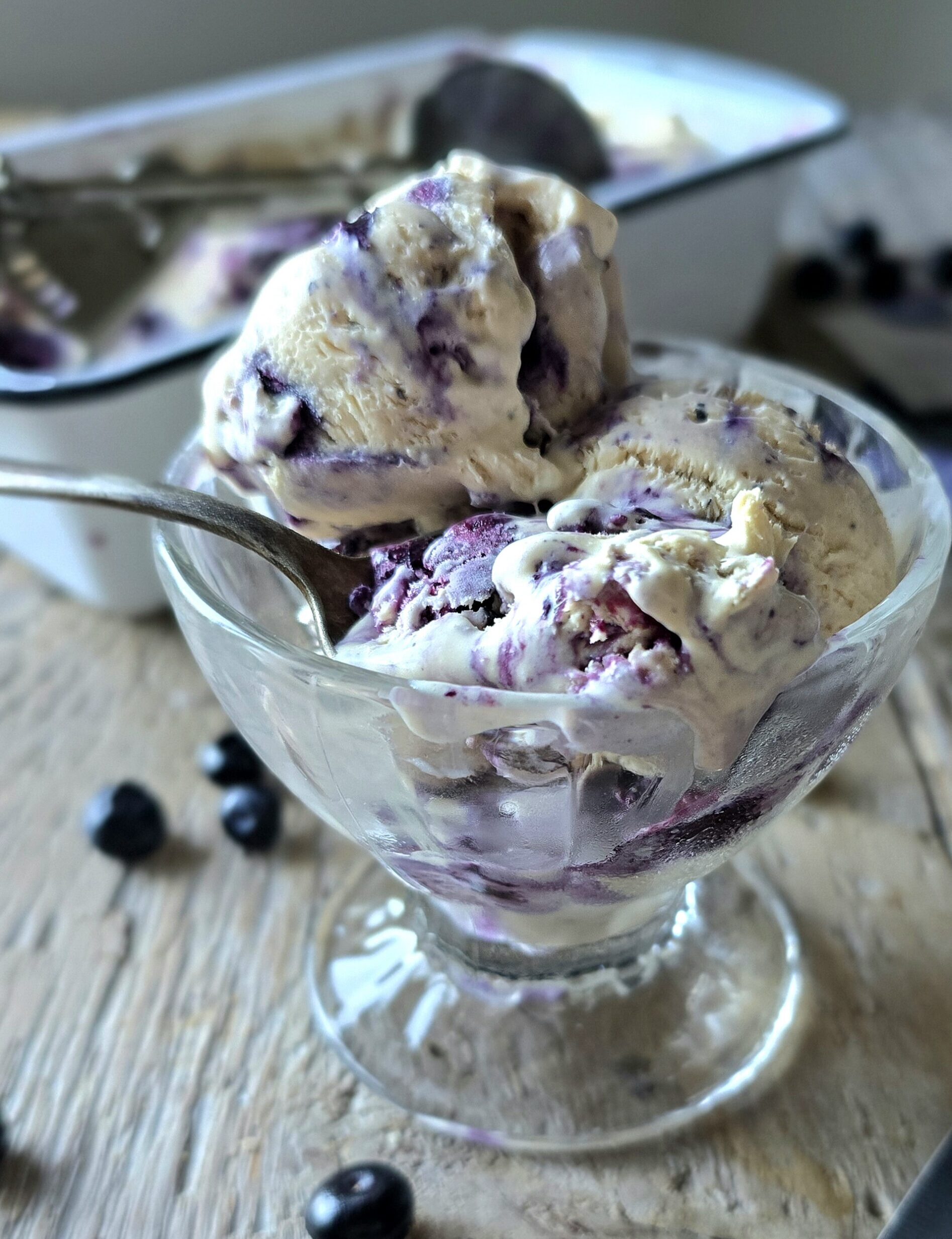 A spoon with melting Earl Grey Ice Cream with Blueberry Lavender Swirl sits in a glass cup.