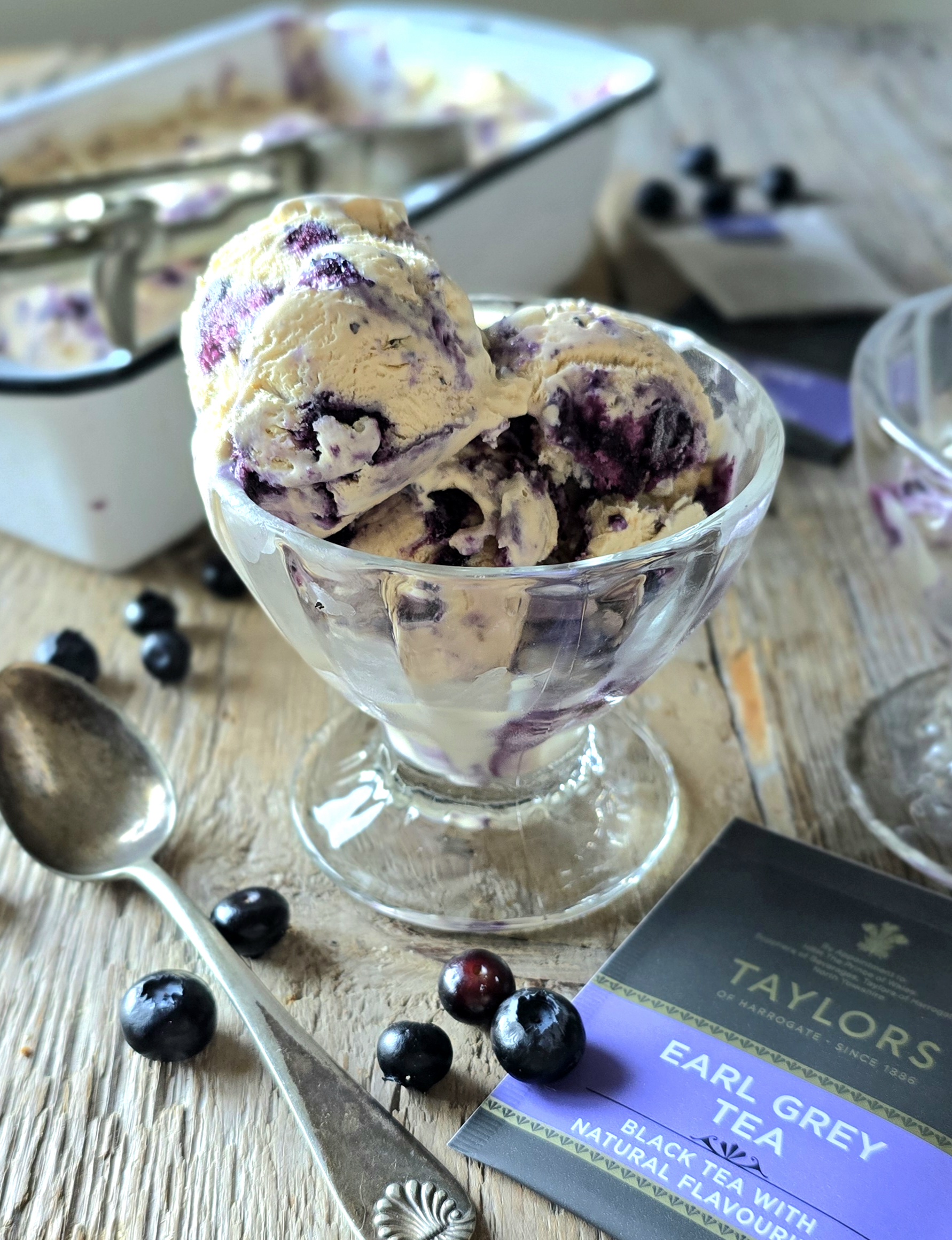 Earl Grey Ice Cream with Blueberry Lavender Swirl is served up in a glass bowl surrounded by fresh blueberries and an earl grey tea bag.