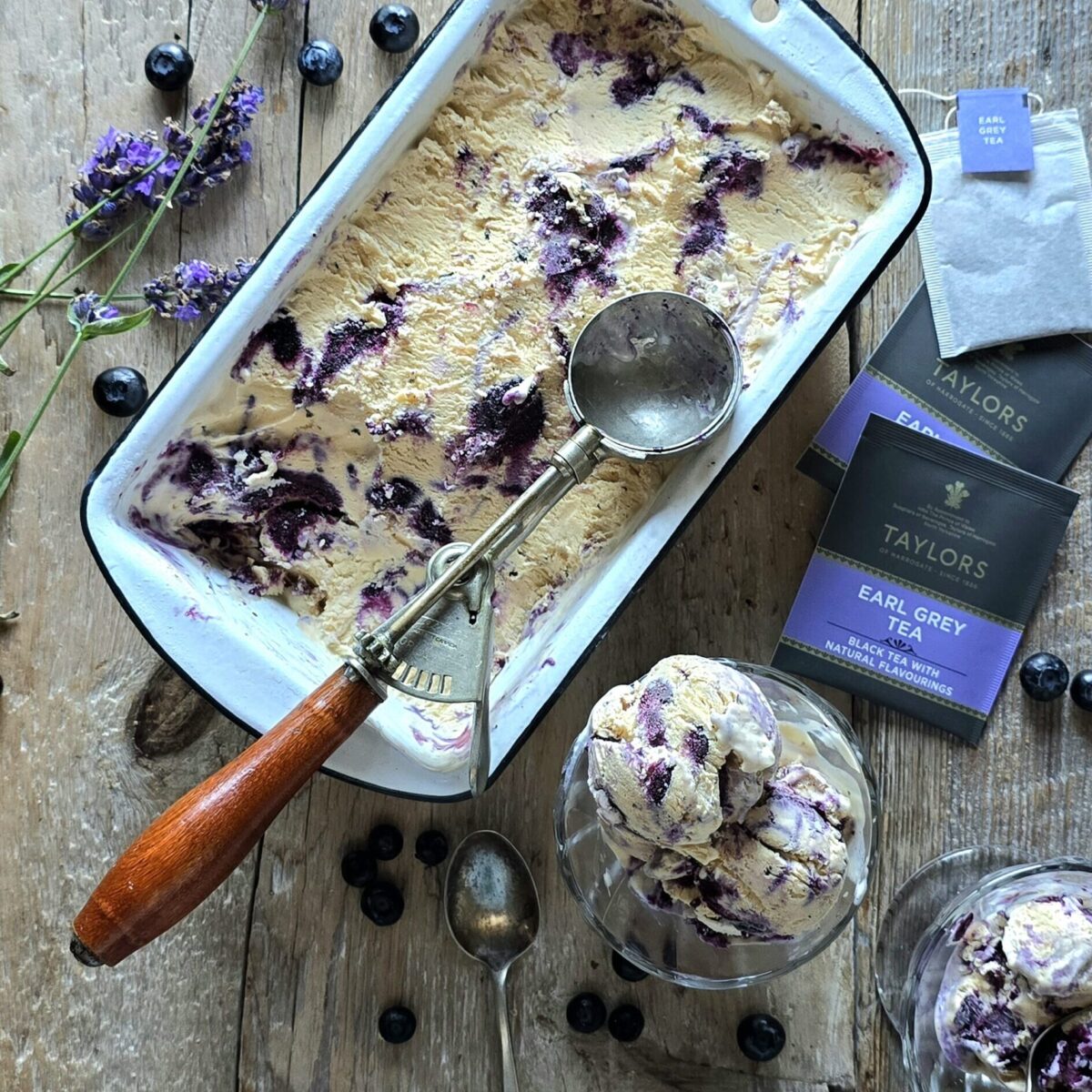 A container of Earl Grey Ice Cream with Blueberry Lavender Swirl sits on the table, with two servings in small cups along side, and fresh lavender and blueberries scattered about.