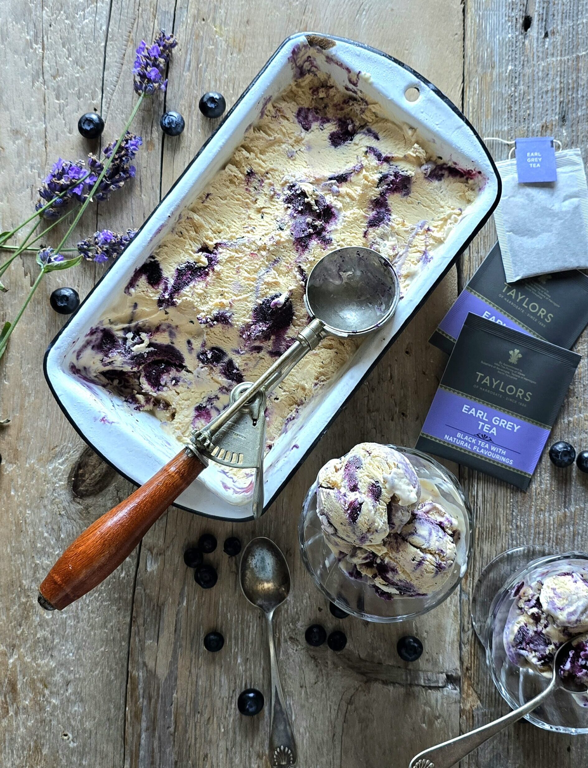 A container of Earl Grey Ice Cream with Blueberry Lavender Swirl sits on the table, with two servings in small cups along side, and fresh lavender and blueberries scattered about.