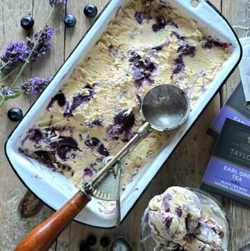 A container of Earl Grey Ice Cream with Blueberry Lavender Swirl sits on the table, with two servings in small cups along side, and fresh lavender and blueberries scattered about.