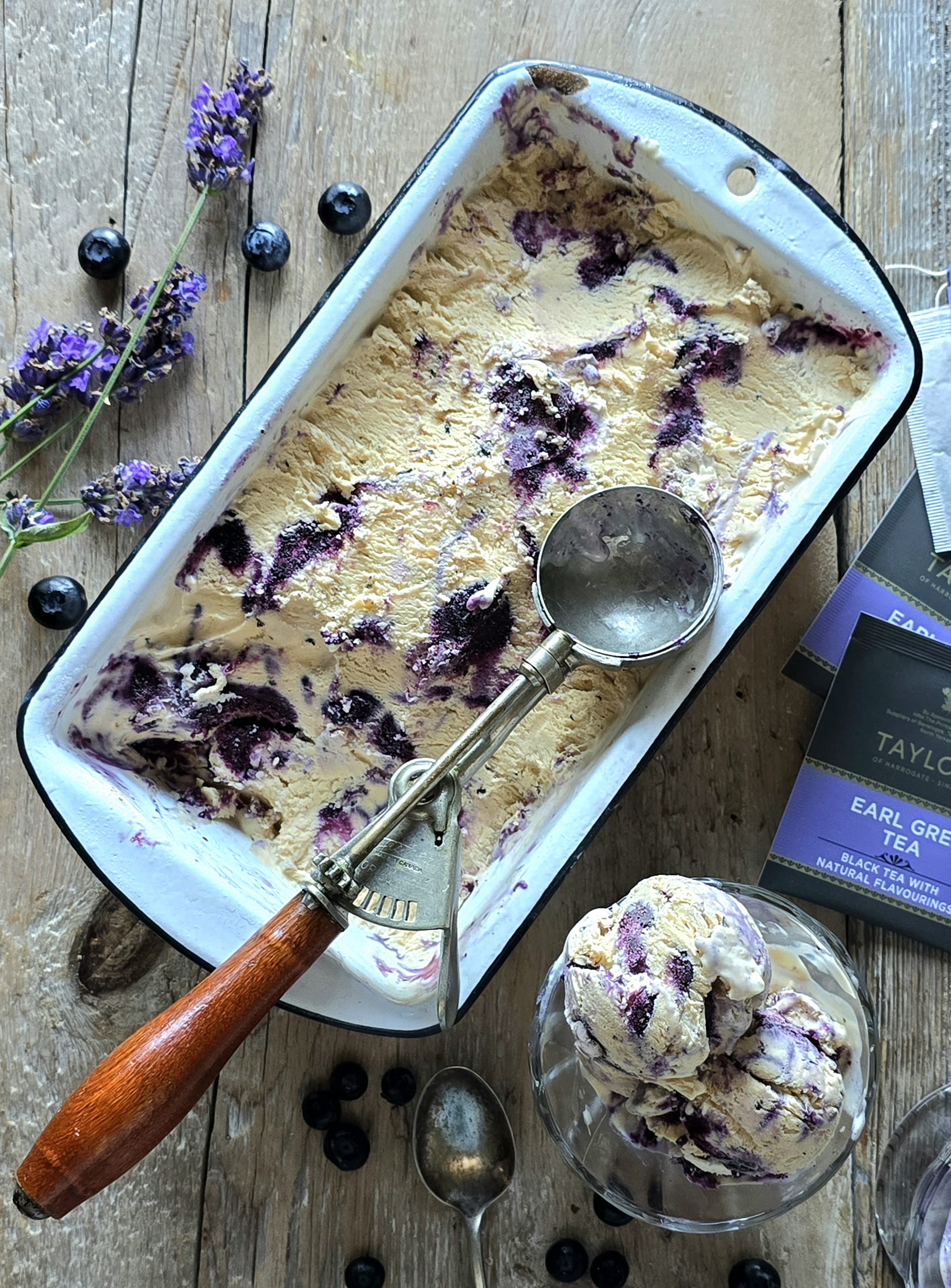 A container of Earl Grey Ice Cream with Blueberry Lavender Swirl sits on the table, with two servings in small cups along side, and fresh lavender and blueberries scattered about.