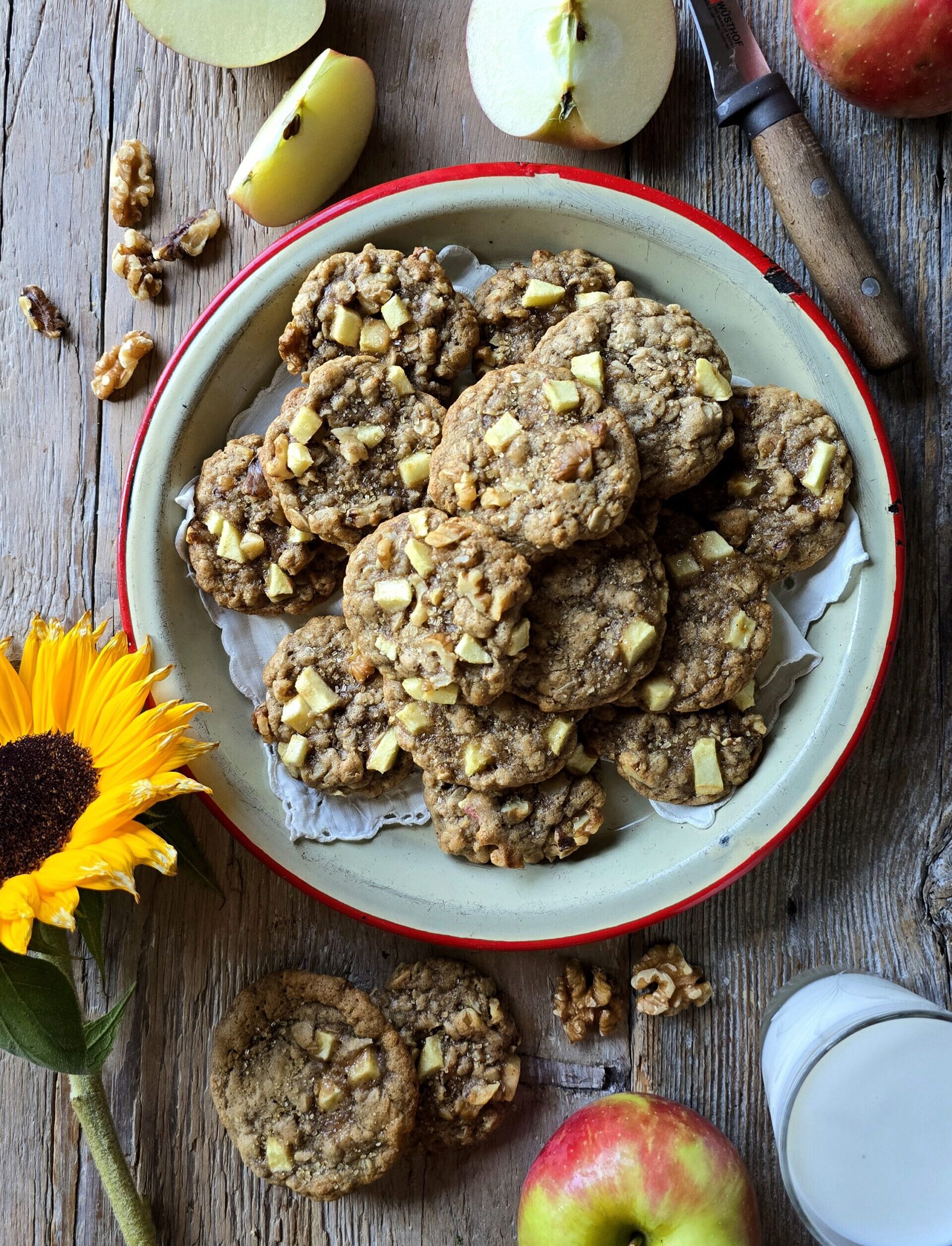 A plate of Chewy Chai Apple Oatmeal cookies is surrounded by fresh cut apples and a sunflower.