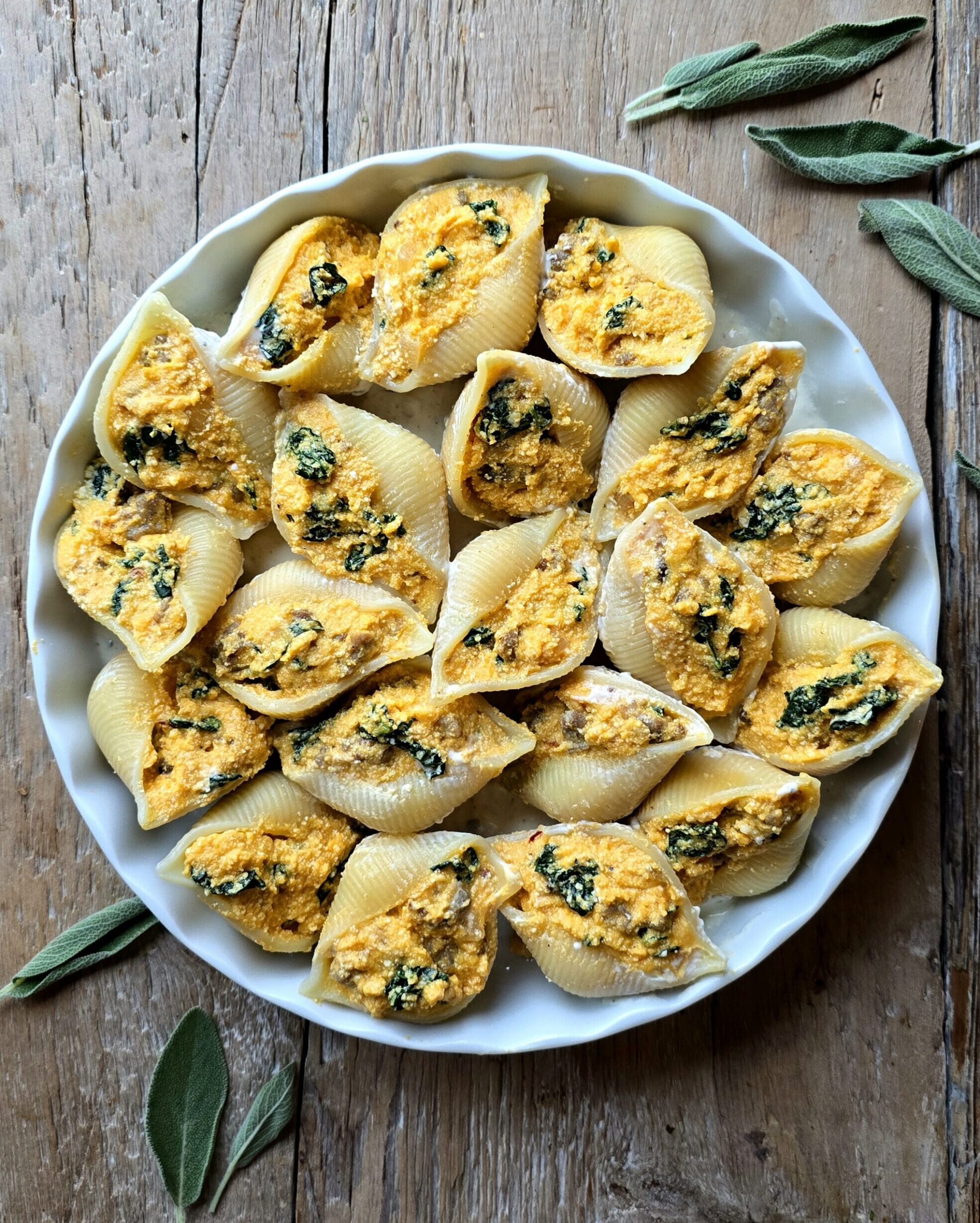 A baking dish filled with Squash, Kale and Sausage Stuffed Pasta Shells sits on the counter with sage leaves to the side.