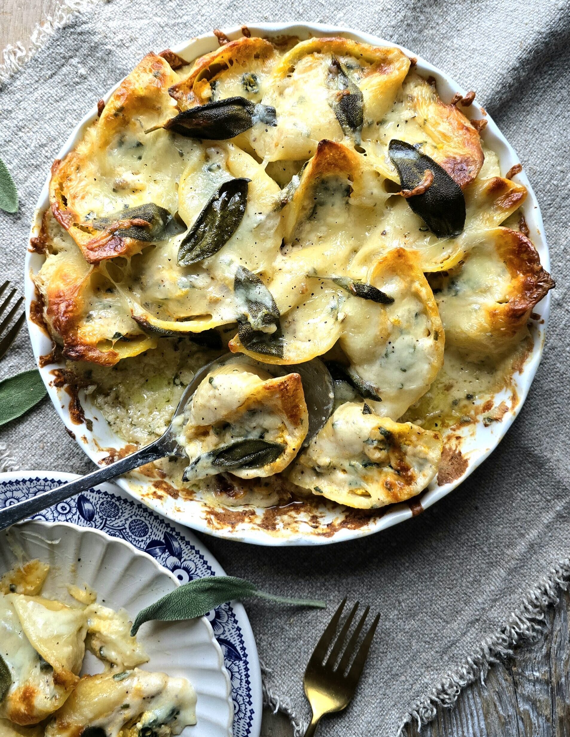 A dish filled with baked Squash, Kale and Sausage Stuffed Shells is being served out, with one plate containing gooey cheese covered shells to the side.