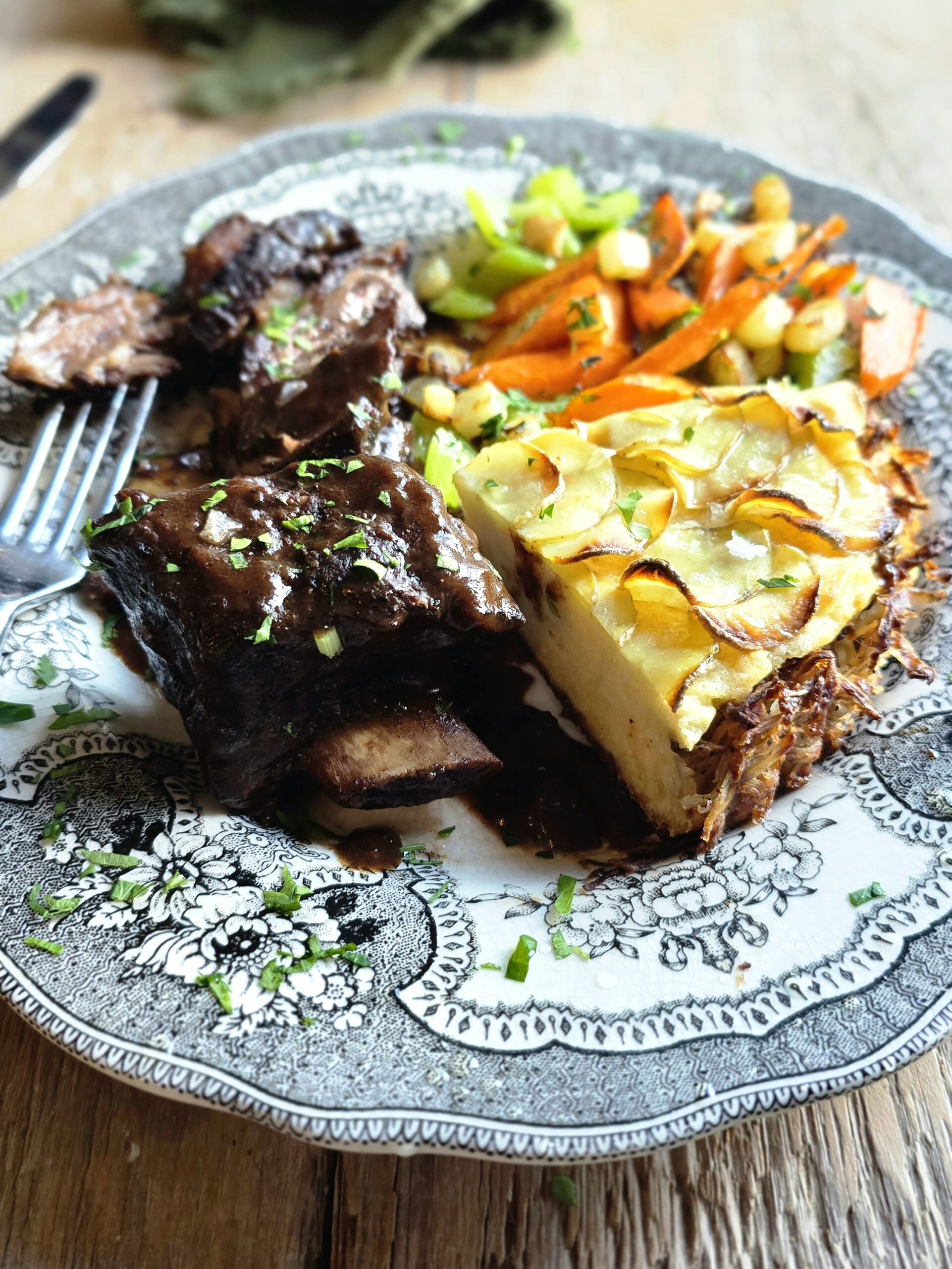 A slice of Potato Pie sits on a plate with braised short ribs and pan roasted vegetables.