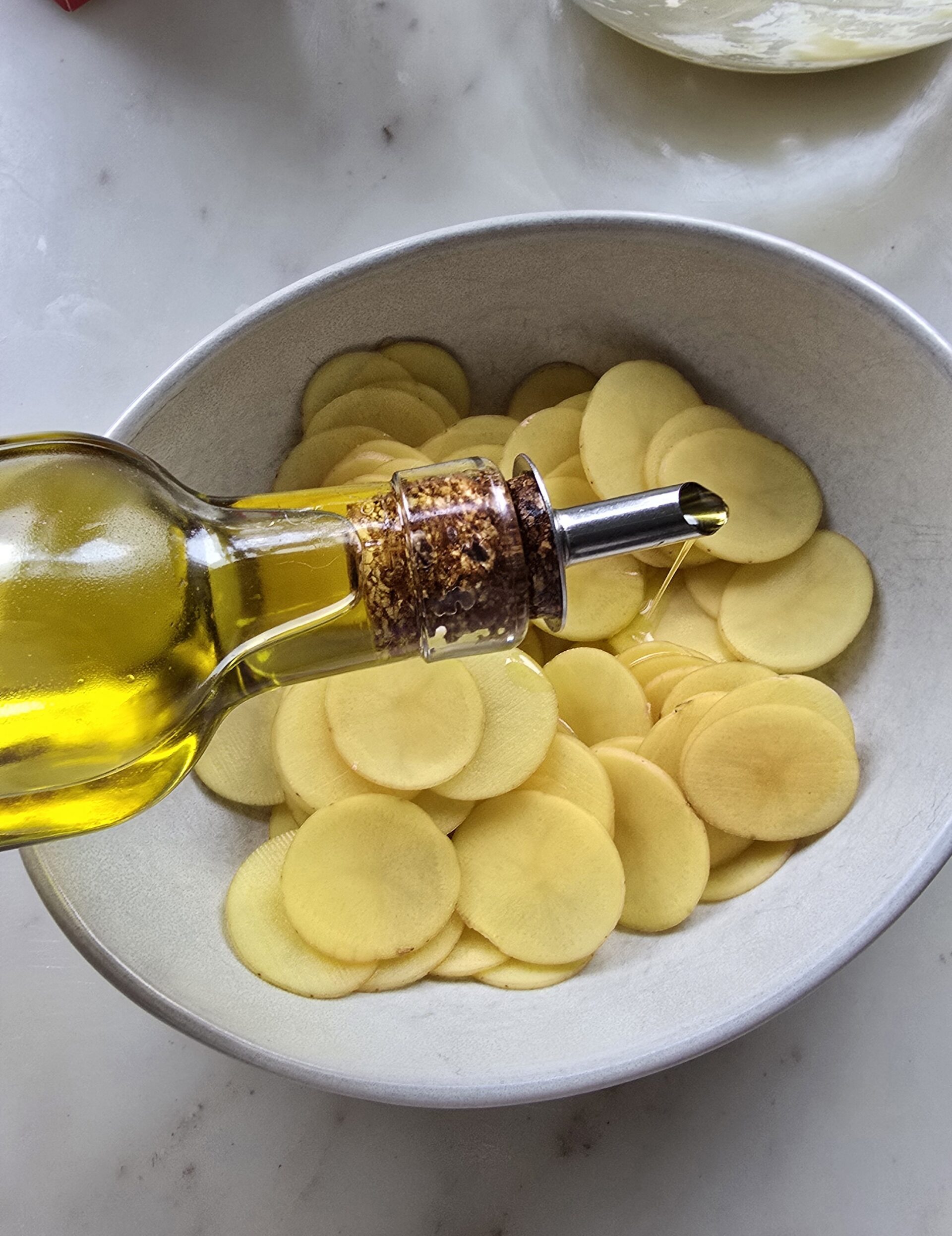 Drizzling olive oil over thinly sliced fingerling potatoes sitting in a bowl.