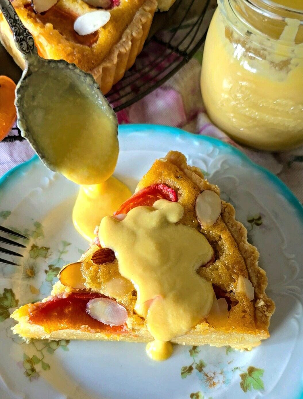 A slice of Apricot Frangipane Tart is on a plate with a spoon pouring creme anglaise over it.