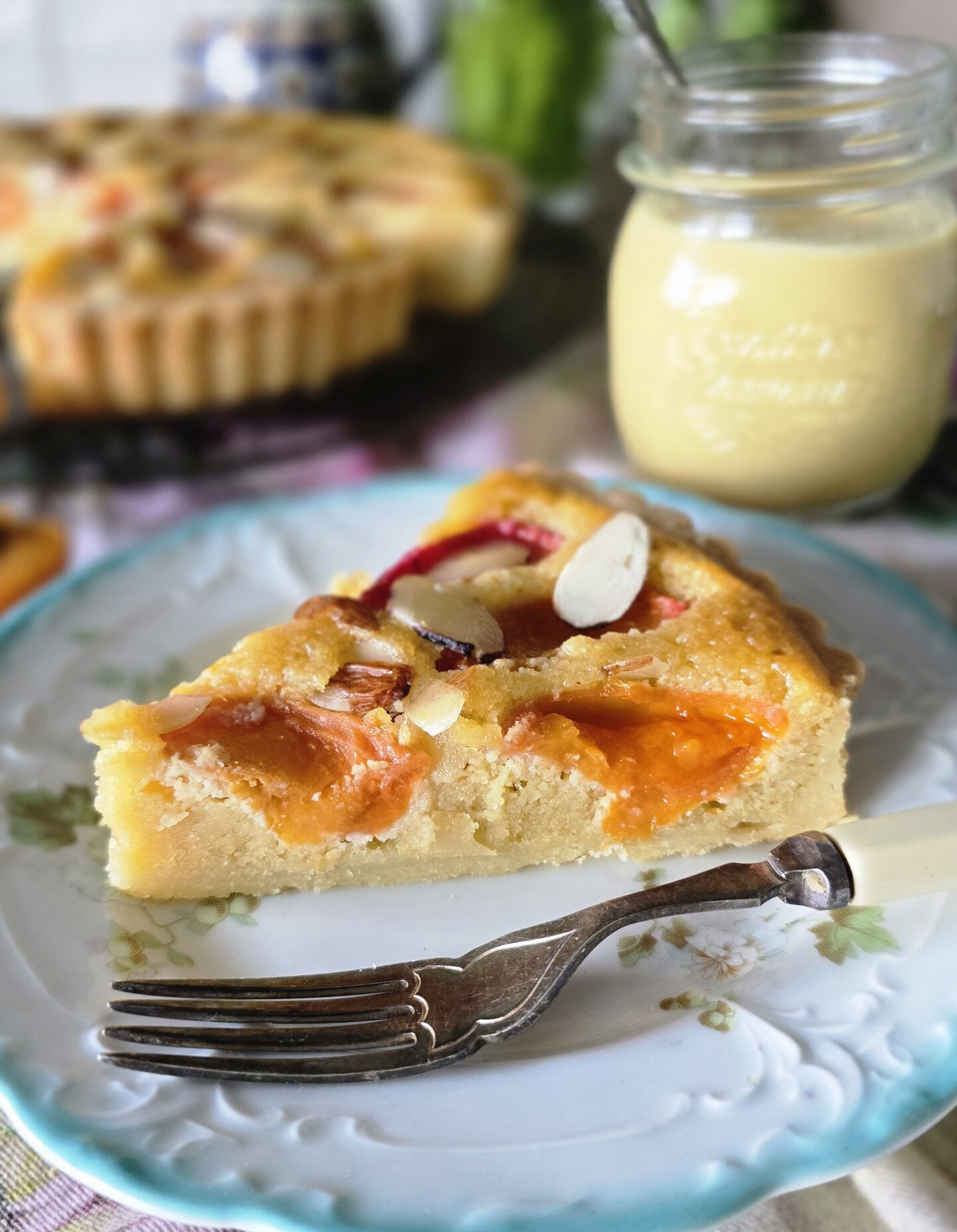 A slice of Apricot Frangipane Tart sits on the counter with a jar of vanilla Creme Anglaise to the side.
