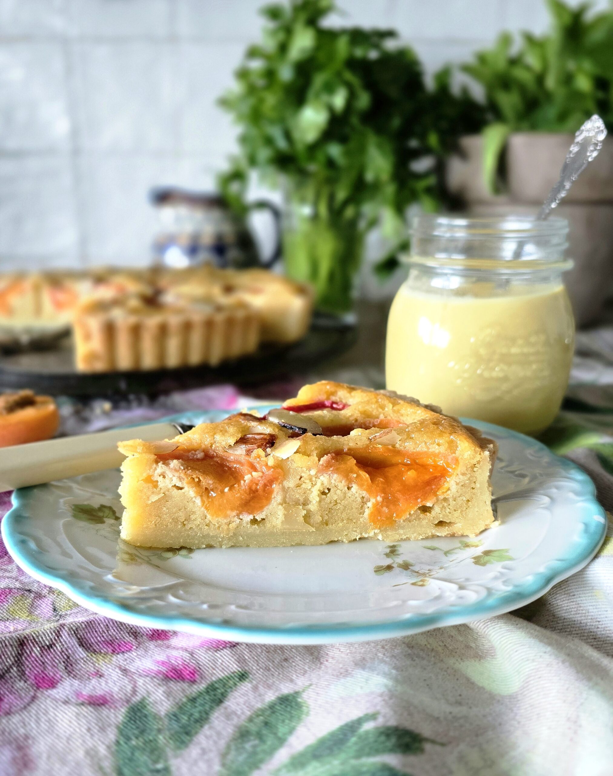 A slice of Apricot Frangipane Tart is on a plate with a jar of freshly made Creme Anglaise in the background.